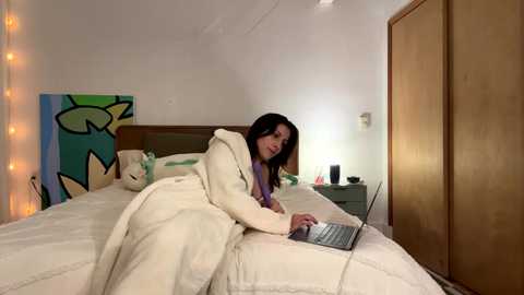 Media: Video of a woman with dark hair, wearing a white robe, lying on a bed with a laptop, in a cozy bedroom with a wooden wardrobe, green wall art, and fairy lights.