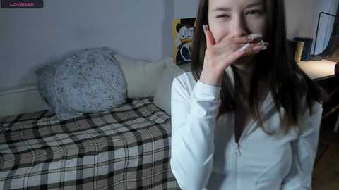 Media: A video of a young woman with long, dark hair, wearing a white zip-up hoodie, blowing a smoke ring. She's in a cozy bedroom with a plaid blanket, floral pillow, and a poster of a cartoon character on the wall.