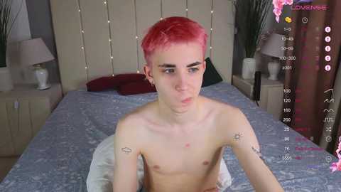 Media: Video of a shirtless young man with short, vibrant pink hair, lying on a bed with a beige headboard and patterned blue sheets. He has small tattoos on his arms and is looking directly at the camera.