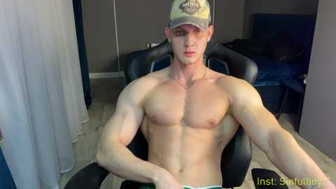 Media: Video of a shirtless, muscular, fair-skinned man in a black gaming chair, wearing a beige baseball cap, in a dimly-lit, modern bedroom with a bed and blue sheets in the background.