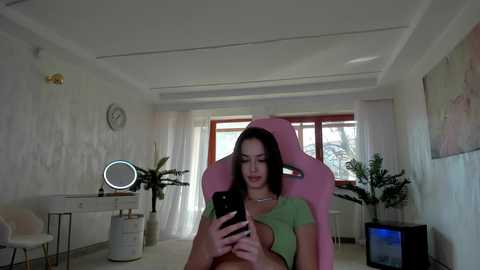 Media: Video of a young woman with fair skin, long dark hair, wearing a green crop top, sitting in a pink gaming chair, in a minimalist, brightly lit room with white walls, a mirror, and plants.