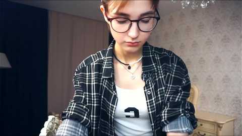 Media: Video of a young woman with glasses, light skin, and brown hair, wearing a black plaid shirt over a white tank top, sitting indoors with beige wallpaper and a wooden nightstand in the background.