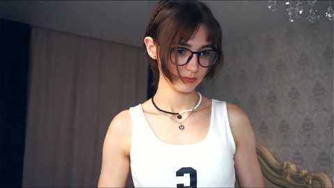 Media: Video of a young, slender Asian woman with short, straight brown hair, wearing black-rimmed glasses, a white tank top, and a necklace, standing in a modern, softly lit room with beige walls and a chandelier.
