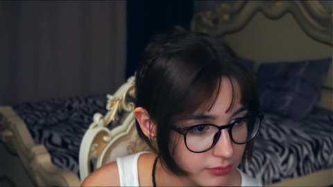 Media: Video of a young woman with dark hair and glasses, wearing a white tank top, lying on a bed with a zebra-patterned comforter in a dimly lit room.
