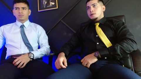Media: Video of two young men in formal attire: one in a light blue shirt, blue tie, and black pants; the other in a black shirt, yellow tie, and black pants, sitting on a black leather chair.