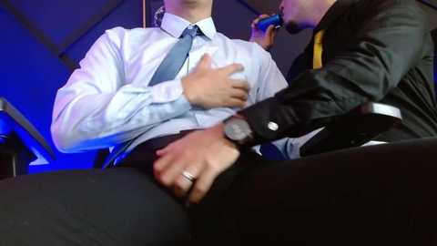 Media: Video of two men in formal attire, one with a light blue shirt and tie, being kissed passionately by another man in a black suit, in a dimly lit, possibly office or private setting.