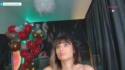 Media: Video of a young woman with straight brown hair and blue eyes, wearing a pink top, looking upward. Background features festive decorations, including red and silver balloons, a heart-shaped display, and dark curtains.
