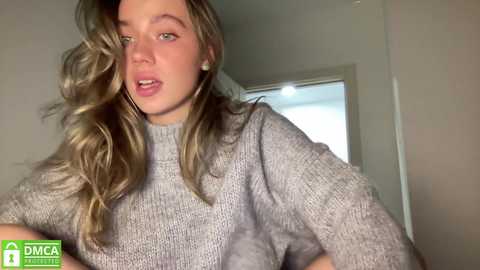 Media: Video of a young Caucasian woman with wavy blonde hair, wearing a grey knitted sweater, leaning against a white wall in a dimly lit room.