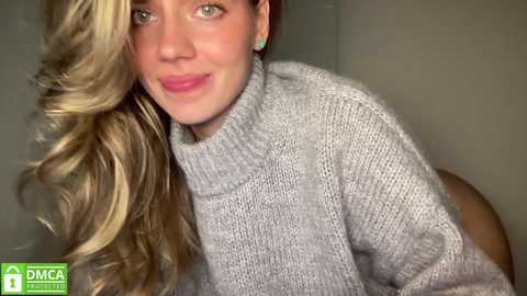 Media: Video of a smiling Caucasian woman with long, wavy blonde hair, wearing a light grey knitted sweater, in a dimly lit room with a DMC logo in the corner.