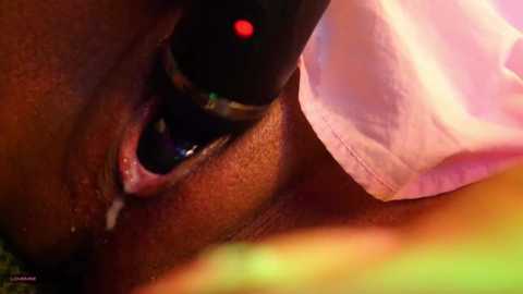 Media: A close-up video of a black woman's eye with a pink, textured contact lens inserted. The image focuses on the eye's iris, pupil, and lens, with a hint of green and yellow background.