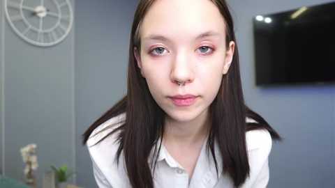 Media: Video of a young, fair-skinned woman with long, straight brown hair, wearing a white button-up shirt, with a septum ring, in a modern room with a clock, TV, and plant.