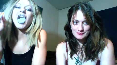 Media: Video of two young women with pale skin and wavy brown hair, one in a black tank top and the other in a white lace top, making exaggerated faces in a dimly lit room.