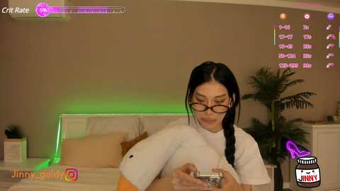 Media: A video of a woman with long black hair, wearing glasses and a white top, holding a phone in a cozy bedroom with a green light, a bed, and a potted plant.