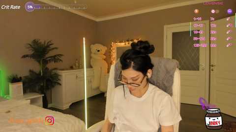 Media: A video of an Asian woman with dark hair in a bun, wearing glasses, and a white shirt, sitting on a chair in a cozy bedroom with a teddy bear, green plants, and dim lighting.