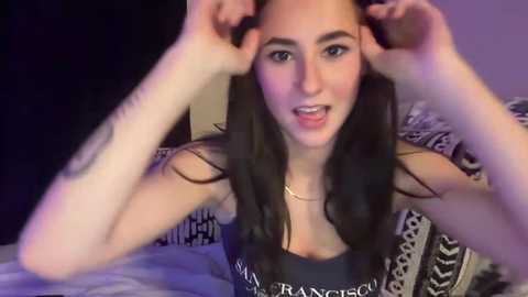 Media: Video of a young woman with long, dark hair, fair skin, and brown eyes, wearing a black tank top with white text, playfully pulling her hair. Background shows a bed with black and white patterned sheets.