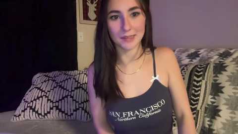 Media: A video of a young woman with long dark hair, fair skin, wearing a black spaghetti strap tank top with \"SF\" and \"NORTH BAY\" text. She is seated on a bed with patterned pillows and a San Francisco-themed blanket.