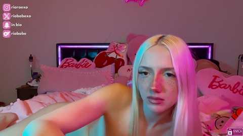 Media: A video of a nude, fair-skinned woman with long, platinum blonde hair lying on a bed adorned with pink \"Barbie\" pillows. The room is dimly lit with pink and purple neon lights.