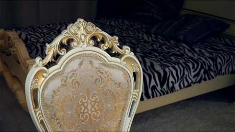 Media: Video of an ornate, gold-framed chair with intricate carvings, positioned in front of a bed with a zebra-patterned black and white bedspread. The background is dimly lit, enhancing the luxurious and vintage aesthetic.