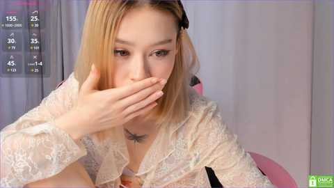 Media: A video of a young Asian woman with long, straight blonde hair, wearing a delicate, lacy white blouse, covering her mouth with her right hand, appearing embarrassed.