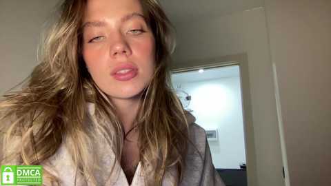 Media: Video of a young Caucasian woman with long, wavy blonde hair, light skin, and full lips, wearing a light grey robe, standing indoors with a blurred background featuring a doorway.