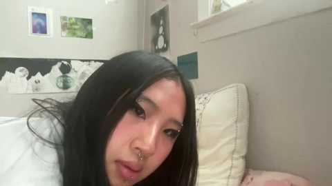 Media: A video of a young Asian woman with long black hair, wearing a septum ring, lying on a bed with white sheets, beige walls, and a black and white abstract art piece on the wall.