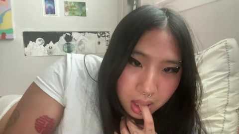 Media: Video of an Asian woman with long black hair, light skin, and a nose ring, wearing a white t-shirt, making a finger heart gesture, in a colorful, graffiti-covered room.