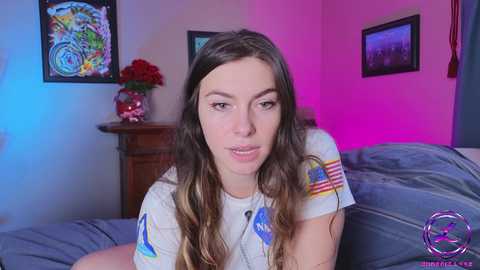 Media: A video of a young woman with long brown hair, wearing a NASA t-shirt, sitting on a bed with a red rose on a nightstand in a pink-lit room.