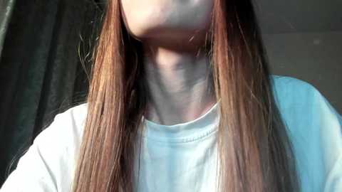 Media: Video of a young woman with long, straight, auburn hair, wearing a white T-shirt, standing against a dark curtain backdrop. Her neck appears elongated and slightly tilted.