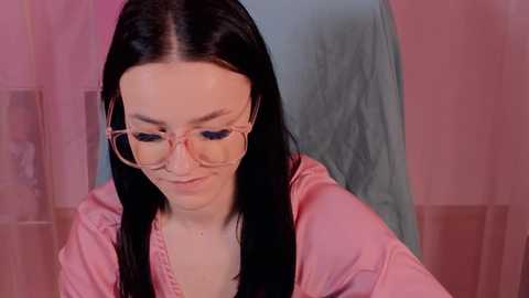 Media: Video of a young woman with fair skin, long black hair, and pink-framed glasses, wearing a pink robe, seated against a pink and blue backdrop.
