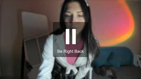 Media: Video of a woman with long black hair, wearing a white cardigan over a pink shirt, sitting on a bed with a blue blanket, in a dimly lit room with a rainbow light effect. Text overlay: \"Be Right Back.\