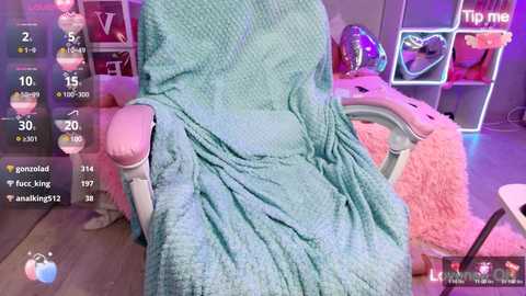 Media: Video of a cozy, plush pink chair draped in a turquoise blanket, with a digital overlay showing live streaming stats. Background features a neon-lit, pink-themed room with shelves and toys.