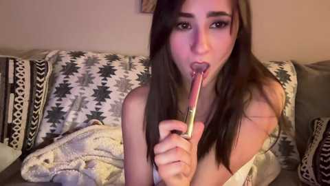 Media: Video of a young woman with long dark hair, light skin, and a slender physique, lying on a patterned couch, eating a popsicle.