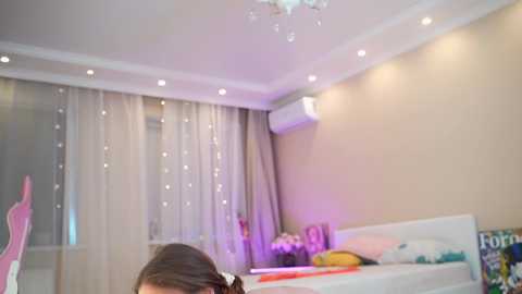 Media: A video of a modern, well-lit bedroom with beige walls, white ceiling, and recessed lights. A person with dark hair is partially visible in the foreground. The room features a white bed with a yellow pillow, and a window with sheer curtains and fairy lights.