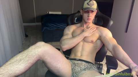 Media: Video of a muscular, shirtless, fair-skinned man with short hair, wearing leopard-print briefs, sitting in a gaming chair in a dimly-lit room with gray walls and a bed.