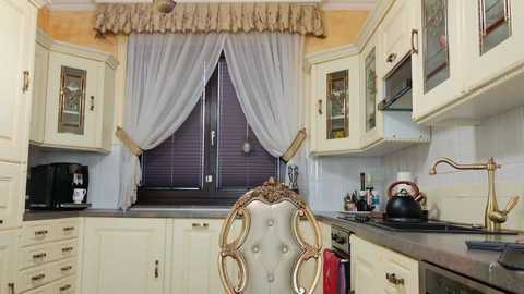 Media: Video of a vintage kitchen with cream cabinets, dark countertops, and a tufted cream chair; features a purple window with sheer curtains, brass fixtures, and a red kettle on the stove.