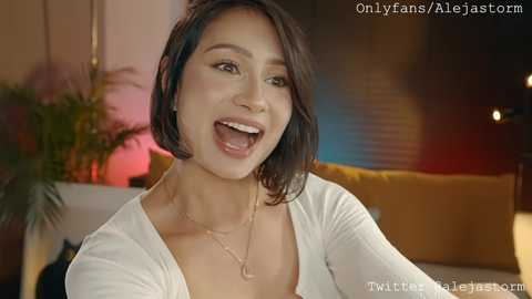 Media: Video of a smiling, light-skinned woman with short black hair, wearing a white top and necklace, indoors with blurred background, OnlyFans watermark.