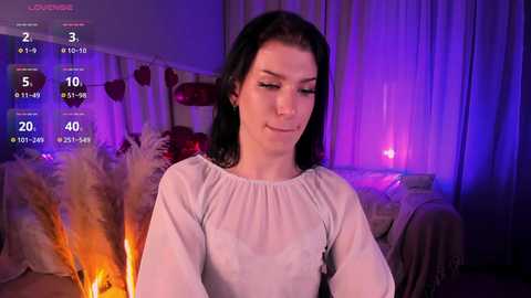 Media: Video of a fair-skinned woman with long black hair, wearing a white blouse, standing in a dimly lit room with purple and orange lighting, surrounded by decorative items like heart-shaped pillows and dried pampas grass.