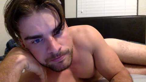 Media: A video of a shirtless man with short dark hair, blue eyes, and a beard, lying on his side on a bed with a dark headboard, looking slightly to the left.