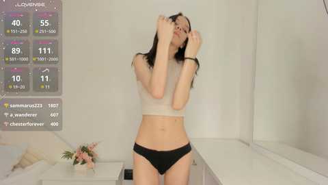 Media: A young woman with fair skin and dark hair stands in a white room, wearing a white crop top and black panties, with digital health metrics displayed on the left.