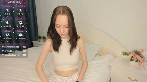 Media: A video of a young, slim, Caucasian woman with shoulder-length brown hair, wearing a white crop top and patterned shorts, sitting on a white bed in a simple, brightly-lit bedroom.