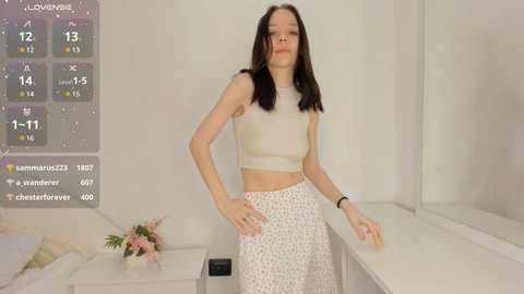 Media: Video of a young woman with straight black hair, wearing a white crop top and floral skirt, standing in a minimalist white room with a calendar overlay.