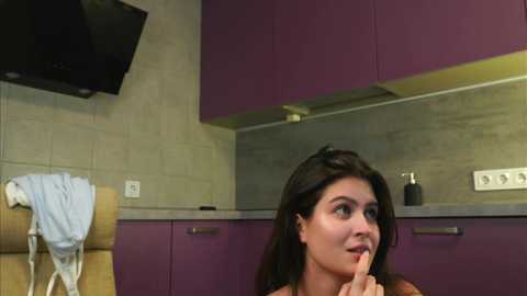 Media: Video of a young woman with dark hair, holding a toothbrush, in a modern kitchen with purple cabinets, a beige wall, and a microwave.