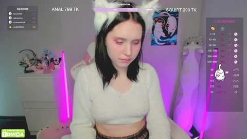 Media: Video of a pale-skinned woman with long black hair wearing white cat ears and a white cropped sweater. She sits in a dimly lit room with neon purple lighting and anime posters on the walls.