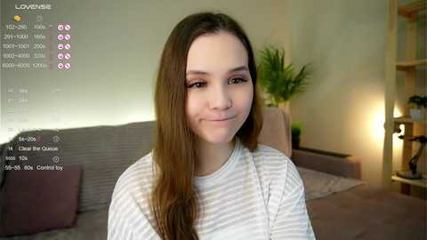Media: Video of a young woman with long brown hair, wearing a striped shirt, smiling. Background shows a living room with a grey couch, a green plant, and a wooden shelf.
