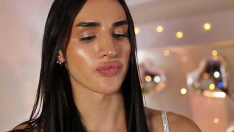 Video of a young woman with long, straight black hair and smooth, sun-kissed skin. She puckers her lips, wearing a silver dress with delicate straps. Background features blurred, warm-toned lights.