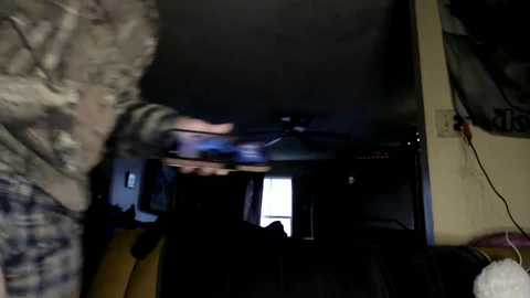 Media: A blurry video showing a person in a camo jacket and another person in a black hoodie, both inside a dimly lit room with a dark TV screen, a laptop, and a few scattered items.