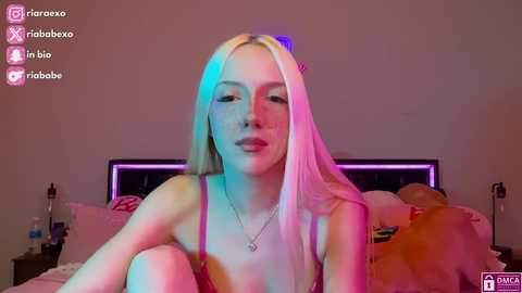 Media: Video of a young, fair-skinned woman with long, platinum blonde hair, wearing a pink bra, sitting on a bed with plush toys. The room is dimly lit with purple and pink neon lights.