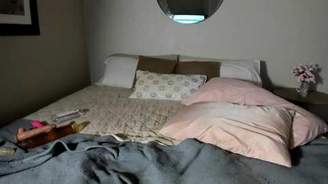 Media: A video of a cluttered bedroom with a bed covered in light pink and beige bedding, surrounded by pillows, a round window, and a small nightstand with a vase of flowers.