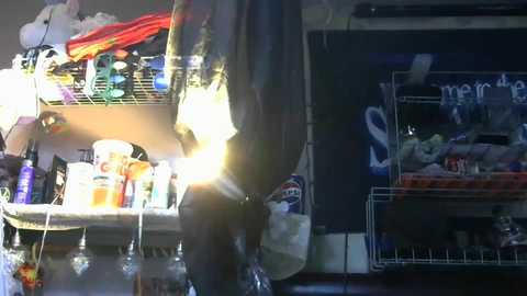 Media: Video of a cluttered kitchen shelf with a hanging black jacket, assorted bottles, and a white box. The background shows a dark cupboard with \"mercy\" written on it.