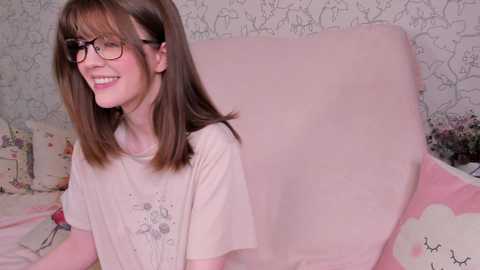 Media: A video of a smiling, fair-skinned, long-haired woman with glasses, wearing a white T-shirt, sitting on a pink bed with a floral-patterned pillow.